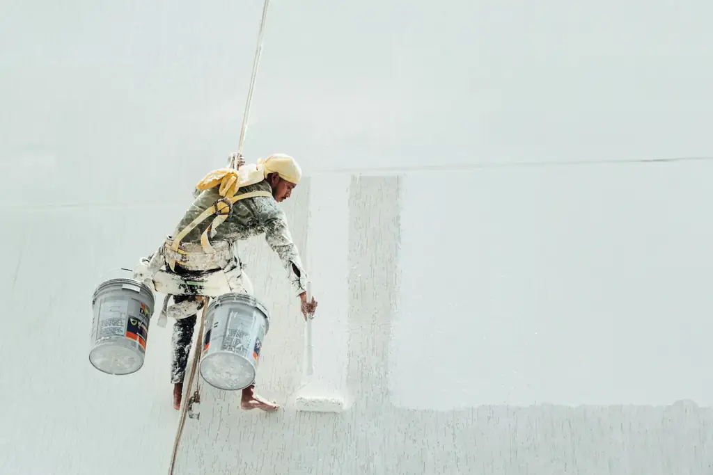 Peintre suspendu le long d’un grand mur blanc, seaux de peinture à la main
